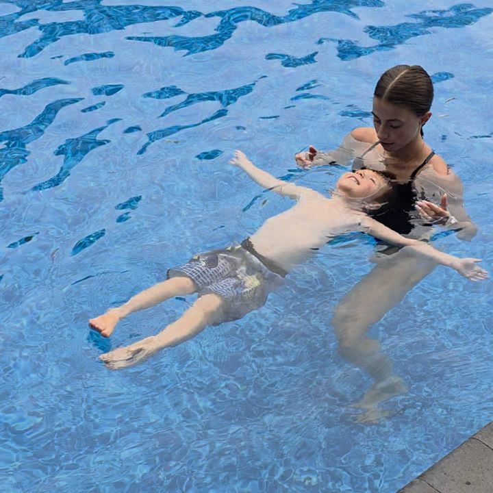 A young swimmer floating on his back with his arms out, Annika supporting him from behind at her backyard pool