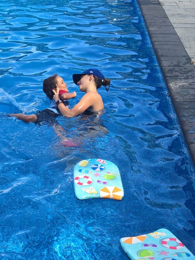Annika holding a smiling toddler in a backyard pool, kickboards floating nearby