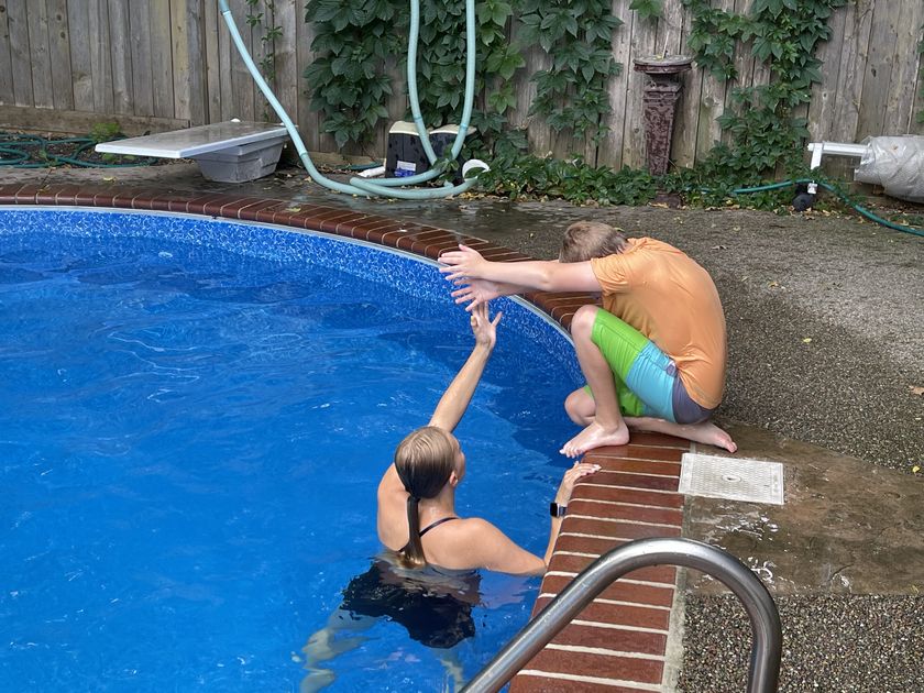 Tayelyn reaching up from the water to a boy sitting on the pool edge, ready to help him in